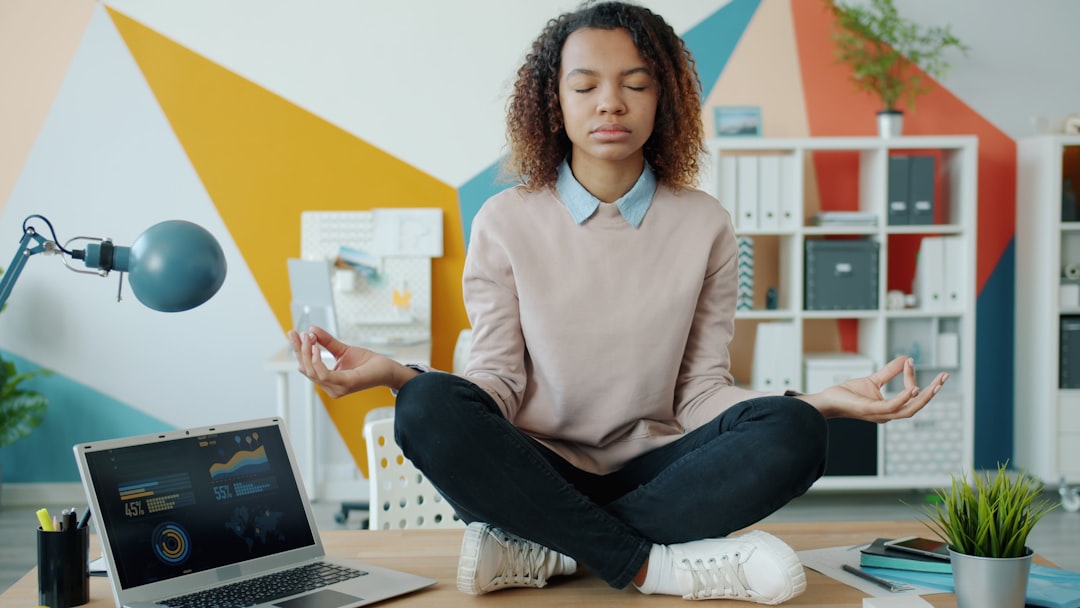 Young African American girl is sitting in lotus pose on office table enjoying relaxation and meditation. Job, stress management and break at work concept.